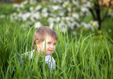 Şık bir tulum giymiş, mavi gözlü sevimli bir çocuk komik bir şekilde gülümsüyor ve uzun yeşil çimenlerin arasına saklanıyor.