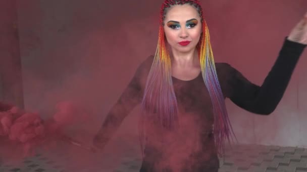 Дівчина в простій чорній сукні з кольоровими косами і веселковим макіяжем позує в червоному товстому димі в теплому весняному місті — стокове відео
