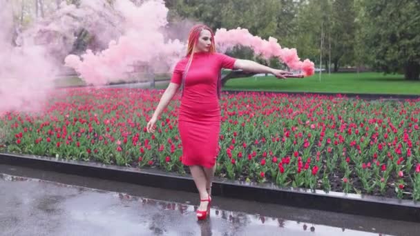 Ein Mädchen mit Make-up mit Regenbogenzöpfen in rotem Kleid posiert in rotem Rauch vor dem Hintergrund eines blühenden Blumenbeetes — Stockvideo