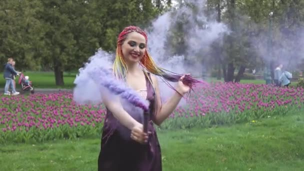 Una chica con una sonrisa con maquillaje brillante y trenzas de colores en un vestido púrpura sopla humo púrpura artificial en un parque — Vídeos de Stock