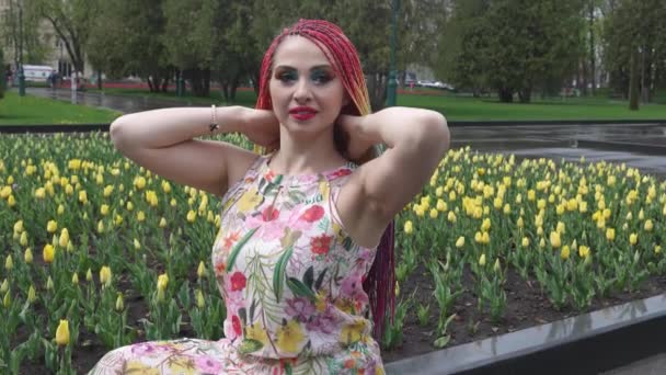 Dziewczyna z jasnym makijażem i afrykańskich tęczowych warkoczach cieszy się nadejściem wiosny w kwitnącym parku w ciepłą pogodę — Wideo stockowe
