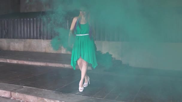 Linda chica en un vestido verde con trenzas africanas y maquillaje colorido posando con humo verde brillante en las calles — Vídeo de stock