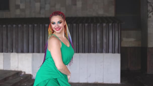 Ένα κορίτσι με μακιγιάζ και πολύχρωμες αφρικανικές κοτσίδες σε ένα πράσινο φόρεμα χαμογελάει έντονα περπατώντας στους δρόμους — Αρχείο Βίντεο