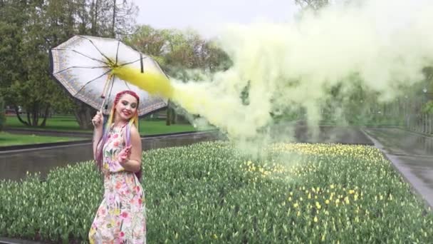 Flicka med afrikanska flätor och smink i vårklänning dansar i regnet med paraply i gul rök i en stadspark — Stockvideo