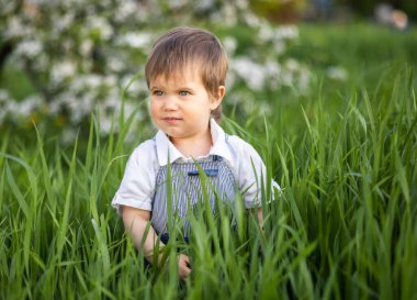 Şık bir tulum giymiş, mavi gözlü sevimli bir çocuk komik bir şekilde gülümsüyor ve uzun yeşil çimenlerin arasına saklanıyor.