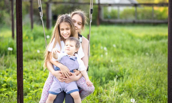 Crianças alegres em roupas leves montam um balanço de rua em um jardim de casa verde florescente e riem em um sorriso feliz — Fotografia de Stock