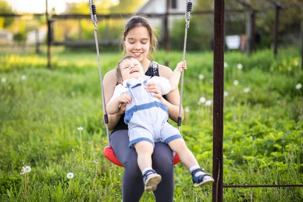 Söt bror och äldre syster rida på en ljus gunga på varm vårdag mot bakgrund av en grön blommande trädgård — Stockfoto