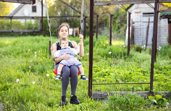 Lindo hermano y hermana mayor paseo en un brillante columpio en cálido día de primavera contra el telón de fondo de un verde jardín en flor —  Fotos de Stock