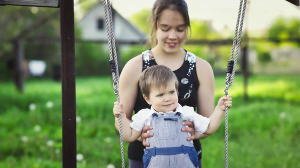 Söt bror och äldre syster rida på en ljus gunga på varm vårdag mot bakgrund av en grön blommande trädgård — Stockfoto