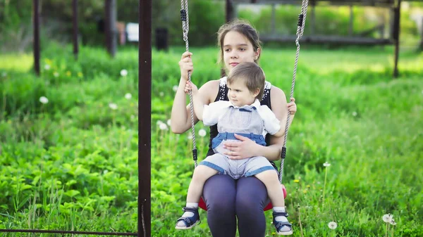 Frate drăguț și sora mai mare plimbare pe un leagăn luminos în ziua de primăvară caldă pe fundalul unei grădini verzi înfloritoare — Fotografie, imagine de stoc