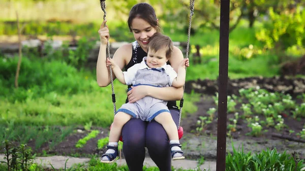 Frate drăguț și sora mai mare plimbare pe un leagăn luminos în ziua de primăvară caldă pe fundalul unei grădini verzi înfloritoare — Fotografie, imagine de stoc