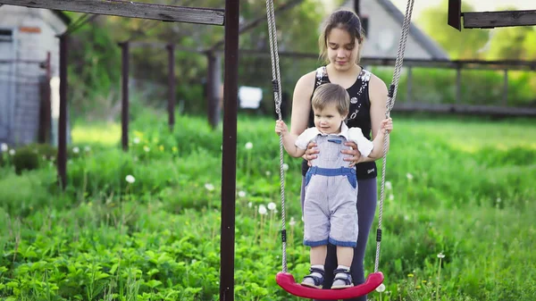 Милий брат і старша сестра катаються на яскравих гойдалках в теплий весняний день на тлі зеленого квітучого саду — стокове фото