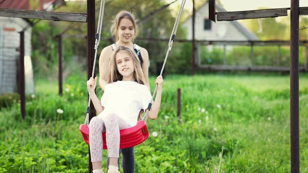 Fröhliche Kinder in leichter Kleidung reiten auf einer Straßenschaukel in einem blühenden, grünen Hausgarten und lachen fröhlich — Stockfoto