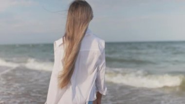 Mavi mayosu ve gömleği olan ince bir kız mavi deniz kıyısında kumlu sahil boyunca yürüyor.