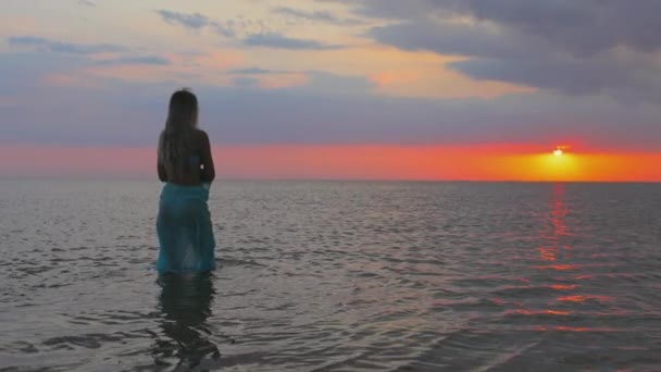 En flicka i blå baddräkt och en ljus pareo poserar mot bakgrunden av en solnedgång i mynningen med transparent vatten — Stockvideo
