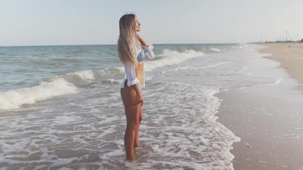 Uma menina delgada em um terno de banho azul suave e camisa caminha ao longo da praia de areia perto do mar azul — Vídeo de Stock