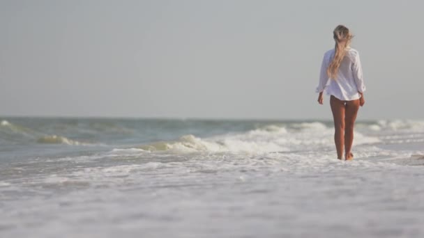 Тонка дівчинка в ніжному блакитному купальнику і сорочці йде по піщаному пляжу біля блакитного моря. — стокове відео