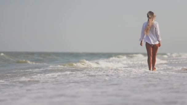 Una ragazza slanciata in un dolce costume da bagno blu e camicia cammina lungo la spiaggia sabbiosa vicino al mare blu — Video Stock