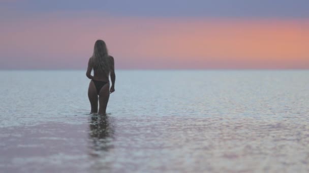 Una chica con el pelo rubio en un traje de baño negro salpica a los lados mientras está sentada en un estuario sobre un fondo al atardecer — Vídeo de stock