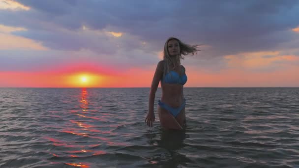 Дівчина з світлим волоссям у блакитному купальнику бризкається з боків, сидячи в лимані на фоні заходу сонця — стокове відео