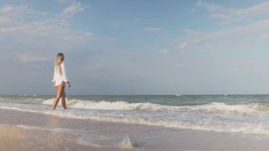 Mavi mayosu ve gömleği olan ince bir kız mavi deniz kıyısında kumlu sahil boyunca yürüyor.