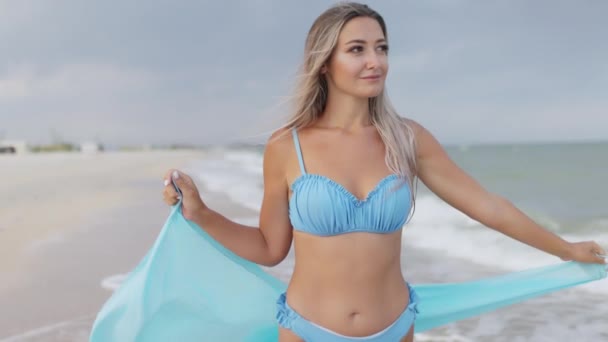 Una ragazza con i capelli biondi in costume da bagno bluastro e uno scialle luminoso passeggia lungo la spiaggia — Video Stock