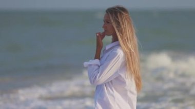 Mavi mayosu ve gömleği olan ince bir kız mavi deniz kıyısında kumlu sahil boyunca yürüyor.