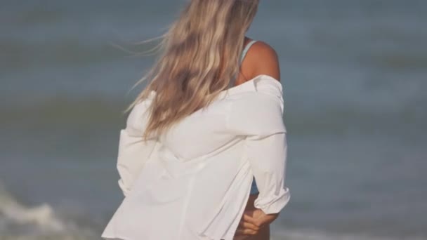 Una ragazza slanciata in un dolce costume da bagno blu e camicia cammina lungo la spiaggia sabbiosa vicino al mare blu — Video Stock