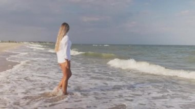 Mavi mayosu ve gömleği olan ince bir kız mavi deniz kıyısında kumlu sahil boyunca yürüyor.