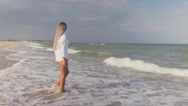 Uma menina delgada em um terno de banho azul suave e camisa caminha ao longo da praia de areia perto do mar azul — Vídeo de Stock