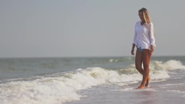 Тонка дівчинка в ніжному блакитному купальнику і сорочці йде по піщаному пляжу біля блакитного моря. — стокове відео