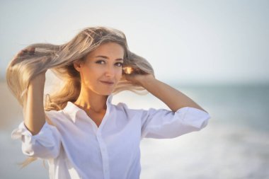 Uzun saçlı, mavi mayolu ve gömlekli bir kız deniz kıyısında saçlarını tarıyor.