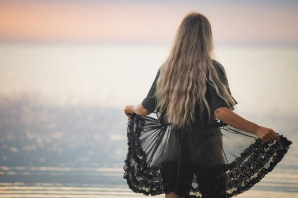 A girl with blond chic hair in a black bikini and a lace shawl walks along the estuary