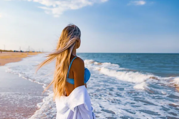Uma menina delgada em um terno de banho azul suave e camisa caminha ao longo da praia de areia perto do mar azul — Fotografia de Stock