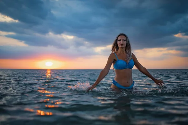 Una chica con el pelo rubio en un traje de baño azul salpica a los lados mientras está sentada en un estuario sobre un fondo al atardecer —  Fotos de Stock