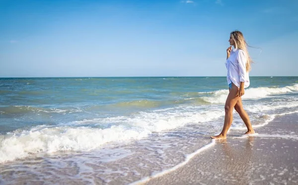 Egy napbarnított lány kék fürdőruhában és világos ingben élvezi a nyarat a tengerparton. — Stock Fotó