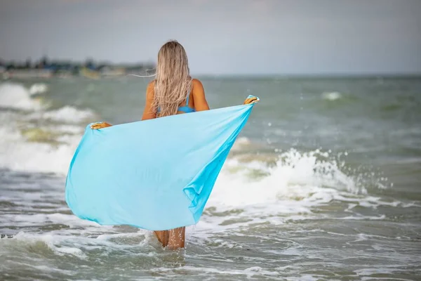 Uma menina com cabelo loiro em um maiô azulado e um xale brilhante caminha ao longo da praia — Fotografia de Stock