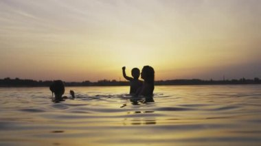 Anne kucağında bebekle kızıyla oynuyor ve gün batımına karşı gölde su sıçratıyor.