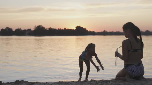 Rolig flicka fångar bubblor på stranden av sjön, som hennes äldre syster släpper — Stockvideo