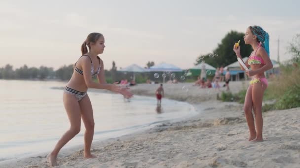 Ung flicka fångar bubblor på stranden av sjön, som hennes lillasyster släpper — Stockvideo