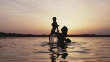 Annem gölde kolsuz çıplak bir bebekle oynuyor yaz günbatımının arka planında.