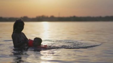 Annem gölde kolsuz çıplak bir bebekle oynuyor yaz günbatımının arka planında.