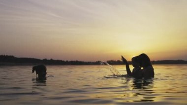 Anne kucağında bebekle kızıyla oynuyor ve gün batımına karşı gölde su sıçratıyor.