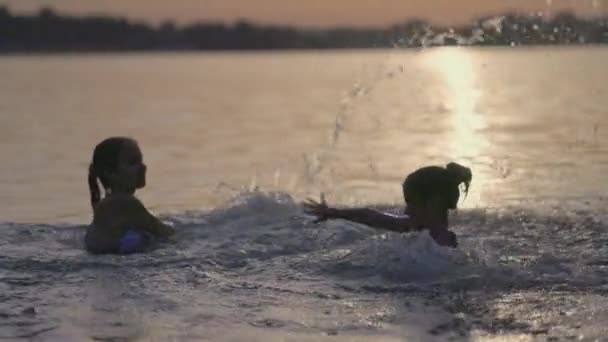 Due sorelle che spruzzano acqua giocando nel lago sullo sfondo del tramonto — Video Stock