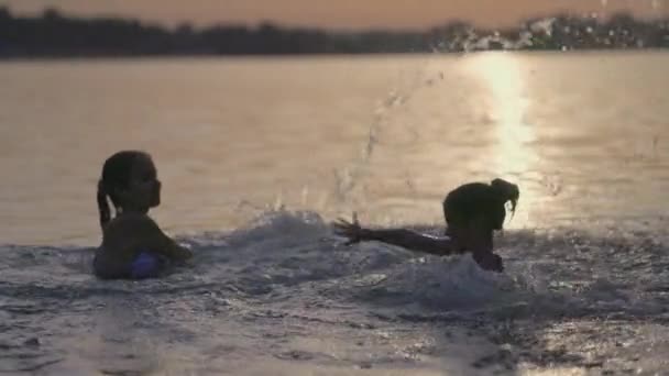 Two sisters splashing water playing in the lake at sunset background — Stock Video