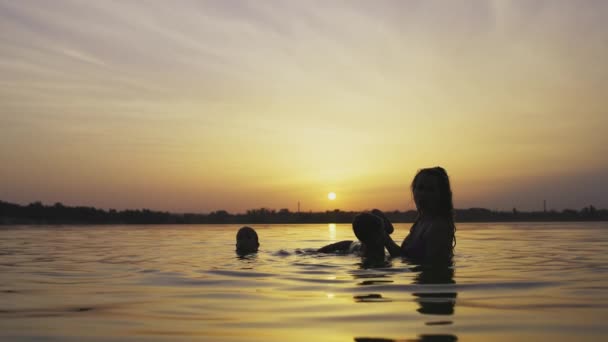 Mãe brinca com um bebê nu em subidas no lago contra o fundo de um pôr do sol de verão — Vídeo de Stock