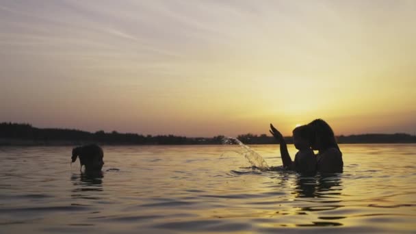 Mamma med barnet hon håller i sina armar leker med sin dotter och stänk i sjön mot bakgrund av solnedgången — Stockvideo