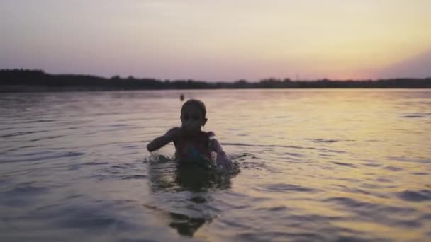 Una chica juega con el agua en el lago y la salpica a los lados contra el telón de fondo de una puesta de sol — Vídeo de stock