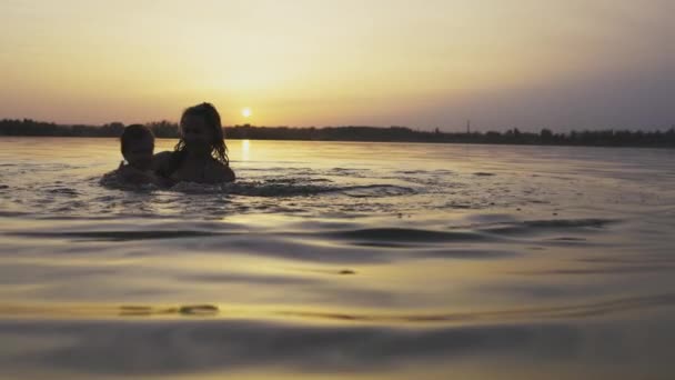 Mãe brinca com um bebê nu em subidas no lago contra o fundo de um pôr do sol de verão — Vídeo de Stock