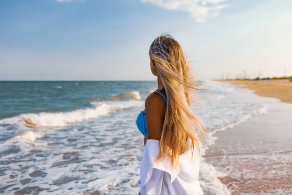 Тонка дівчинка в ніжному блакитному купальнику і сорочці йде по піщаному пляжу біля блакитного моря. — стокове фото